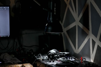 A dimly lit room contains a DJ setup with headphones resting on a mixer. A large lamp extends from the top, casting focused light onto the equipment. The background features a dark wall with geometric patterns and part of a computer monitor is visible on the left.
