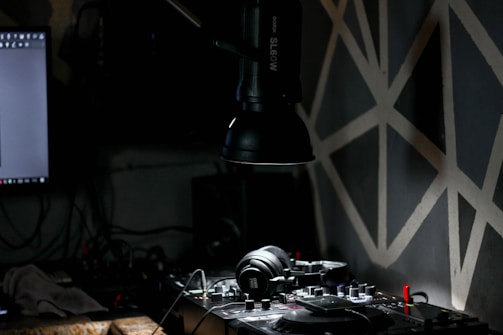 A dimly lit room contains a DJ setup with headphones resting on a mixer. A large lamp extends from the top, casting focused light onto the equipment. The background features a dark wall with geometric patterns and part of a computer monitor is visible on the left.