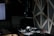 A dimly lit room contains a DJ setup with headphones resting on a mixer. A large lamp extends from the top, casting focused light onto the equipment. The background features a dark wall with geometric patterns and part of a computer monitor is visible on the left.