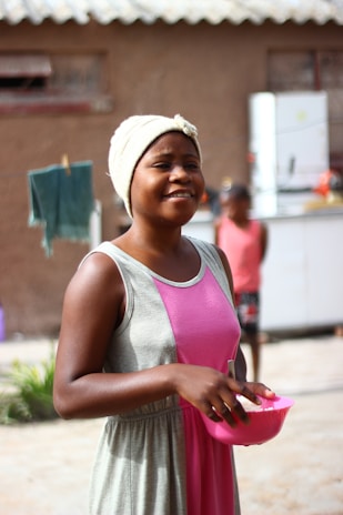 A close-up of a smiling person holding a colorful bowl of whole foods outside on a sunny day.