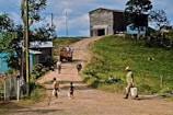A rural scene features a dirt road leading up to a large wooden building on a hill. Several people are walking along the road, including a man with a white bucket and two children holding similar containers. A vehicle with passengers is seen in the background, and a man is guiding a cow. Trees and grassy areas surround the structures, creating a pastoral setting.