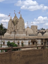 A large, ornate temple with intricate carvings and multiple spires, topped with flags. The temple is surrounded by lush green trees and set against a backdrop of partly cloudy skies. The architecture features a cream or beige color palette with detailed stone work.