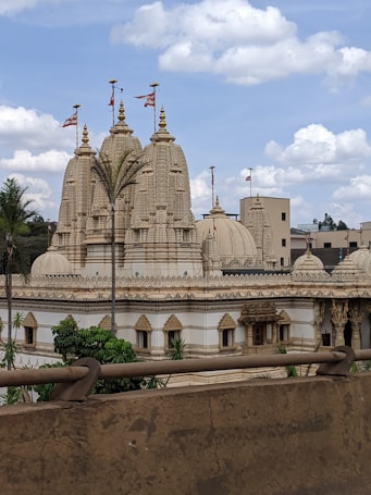 A large, ornate temple with intricate carvings and multiple spires, topped with flags. The temple is surrounded by lush green trees and set against a backdrop of partly cloudy skies. The architecture features a cream or beige color palette with detailed stone work.