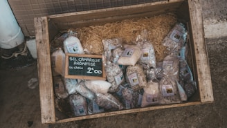 A wooden crate on the ground contains various bags of aromatic salt from Camargue. The bags are neatly arranged on a bed of straw, and there is a small chalkboard sign in the center displaying the product type and price.
