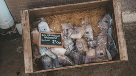 A wooden crate on the ground contains various bags of aromatic salt from Camargue. The bags are neatly arranged on a bed of straw, and there is a small chalkboard sign in the center displaying the product type and price.