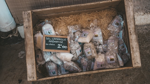A wooden crate on the ground contains various bags of aromatic salt from Camargue. The bags are neatly arranged on a bed of straw, and there is a small chalkboard sign in the center displaying the product type and price.
