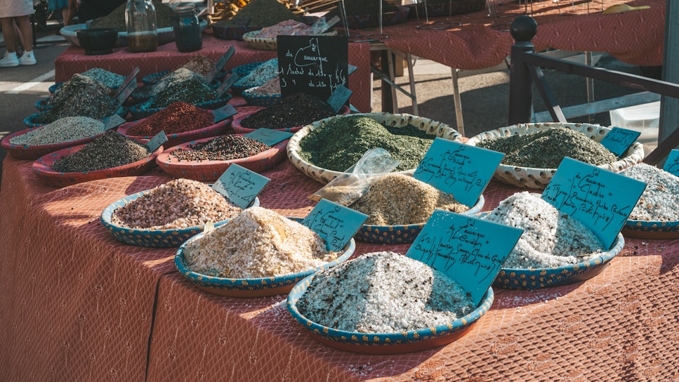 A market stall displays a variety of spices and herbs in shallow bowls, with handwritten labels placed on top of each pile. The spices have different colors and textures, including white, green, black, and pink. The tablecloth beneath the bowls has a geometric pattern, and sunlight casts shadows creating a warm and lively ambiance.