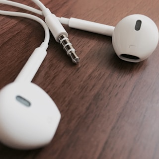White earphones with a 3.5 mm audio jack placed on a wooden surface. The earphones have a smooth, streamlined design and the jack has a metallic finish with several sections.