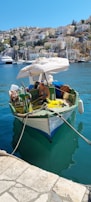 A small fishing boat is docked beside a stone pier in a calm harbor. A man aboard the boat is repairing fishing nets. The boat is equipped with various tools and fishing equipment, and an umbrella provides shade. In the background, a picturesque hillside town with numerous buildings and sailboats gently floating on the water can be seen.