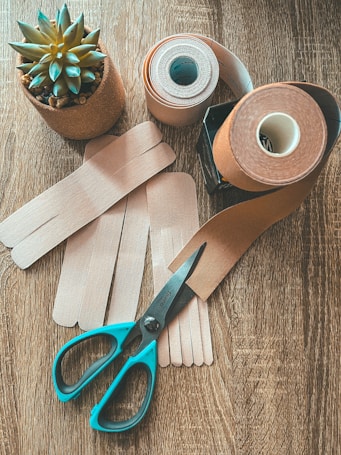 On a wooden surface, a small potted succulent plant sits beside rolls of brown and beige adhesive bandages. A pair of turquoise-handled scissors lies nearby, with some bandage strips laid out flat.