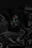 A sleek, dark bottle of perfume sits on an elegantly draped black satin fabric. The smooth texture of the fabric complements the glossy surface of the bottle, which has a metallic cap.