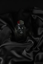 A sleek, dark bottle of perfume sits on an elegantly draped black satin fabric. The smooth texture of the fabric complements the glossy surface of the bottle, which has a metallic cap.