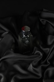 A sleek, dark bottle of perfume sits on an elegantly draped black satin fabric. The smooth texture of the fabric complements the glossy surface of the bottle, which has a metallic cap.