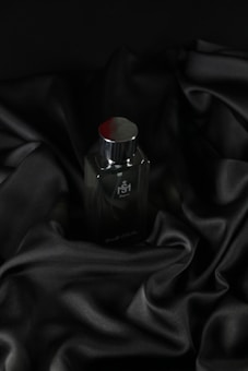 A sleek, dark bottle of perfume sits on an elegantly draped black satin fabric. The smooth texture of the fabric complements the glossy surface of the bottle, which has a metallic cap.