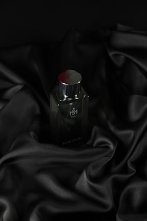 A sleek, dark bottle of perfume sits on an elegantly draped black satin fabric. The smooth texture of the fabric complements the glossy surface of the bottle, which has a metallic cap.