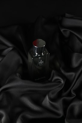 A sleek, dark bottle of perfume sits on an elegantly draped black satin fabric. The smooth texture of the fabric complements the glossy surface of the bottle, which has a metallic cap.