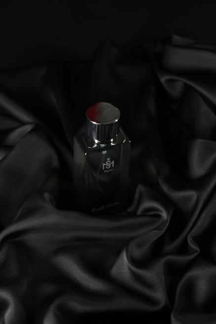A sleek, dark bottle of perfume sits on an elegantly draped black satin fabric. The smooth texture of the fabric complements the glossy surface of the bottle, which has a metallic cap.