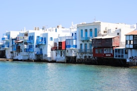 A row of charming waterfront houses with colorful wooden balconies overlooks a clear, blue sea. The buildings feature a mix of white walls and accents in shades of blue and red. The architecture is traditional, characterized by cubic forms and flat roofs, commonly seen in Mediterranean coastal towns.