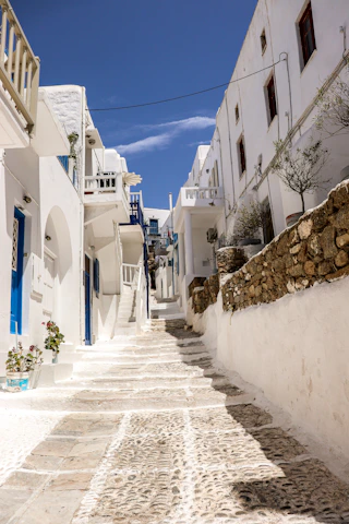 Sunlit cobblestone street in a charming Greek village with whitewashed houses and blue shutters.
