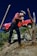 A person wearing a red plaid shirt and blue jeans is mending a fence on a sloped, rocky terrain. The individual is using a tool to secure wooden fence posts. In the background, there is a red vintage truck parked amidst sparse greenery under a clear blue sky.