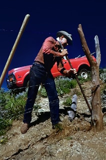 A person wearing a red plaid shirt and blue jeans is mending a fence on a sloped, rocky terrain. The individual is using a tool to secure wooden fence posts. In the background, there is a red vintage truck parked amidst sparse greenery under a clear blue sky.