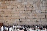A large ancient stone wall with several people gathered in front of it, some wearing religious garments or shawls. The people appear to be engaged in a form of worship or gathering. White plastic chairs and tables are also present, suggesting an organized setting.