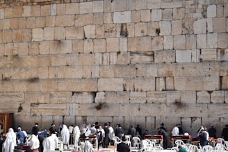 A large ancient stone wall with several people gathered in front of it, some wearing religious garments or shawls. The people appear to be engaged in a form of worship or gathering. White plastic chairs and tables are also present, suggesting an organized setting.