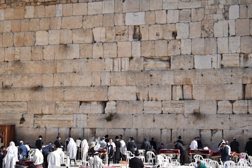 A large ancient stone wall with several people gathered in front of it, some wearing religious garments or shawls. The people appear to be engaged in a form of worship or gathering. White plastic chairs and tables are also present, suggesting an organized setting.