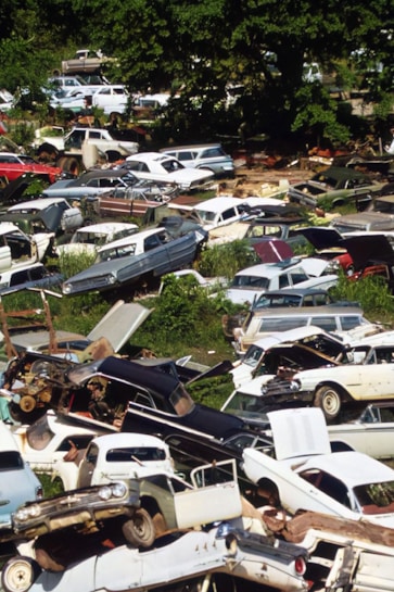 A densely packed junkyard filled with various old and rusted cars. Some vehicles have missing parts like doors and hoods. The cars are stacked haphazardly, surrounded by overgrown grass and trees.