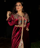 A model wearing a rich crimson dress with vivid terracotta patterns, standing against an ivory white textured wall