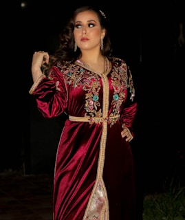 A woman stands in a dark, possibly outdoor setting, wearing a richly decorated velvet red dress adorned with intricate gold and colorful floral embroidery. Her hair is styled in loose curls, and she wears minimal jewelry, including earrings. The expression on her face is calm and poised, looking slightly to the side.