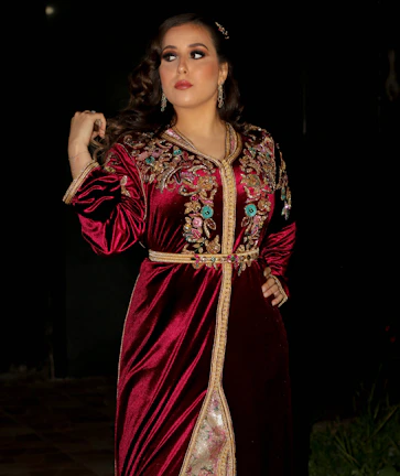 A model wearing a rich crimson dress with vivid terracotta patterns, standing against an ivory white textured wall