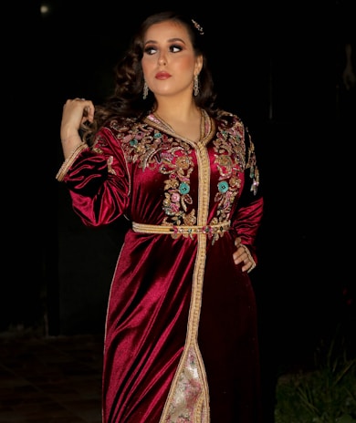 A woman stands in a dark, possibly outdoor setting, wearing a richly decorated velvet red dress adorned with intricate gold and colorful floral embroidery. Her hair is styled in loose curls, and she wears minimal jewelry, including earrings. The expression on her face is calm and poised, looking slightly to the side.