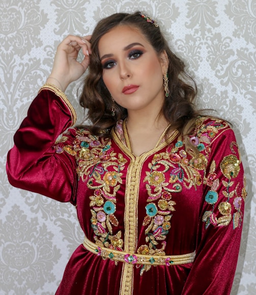 A woman is wearing a richly embroidered maroon dress with gold, pink, green, and blue floral embellishments and intricate designs. Her hair is styled in loose curls, and she is wearing makeup with pronounced eyeshadow and lipstick. The background features a patterned wallpaper.