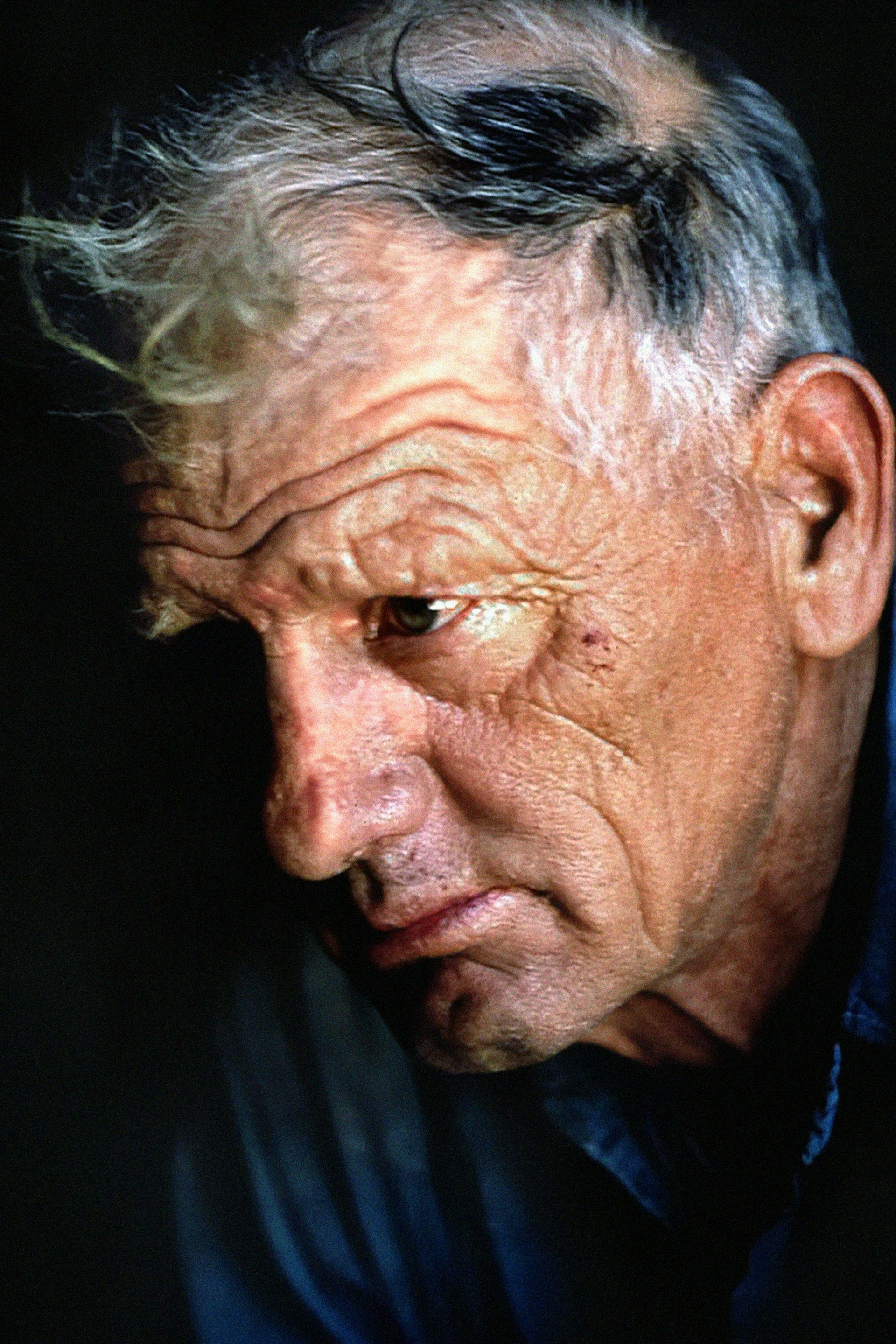 A portrait of an elderly man with a weathered complexion and deep wrinkles, captured in a moment of introspection with a somber expression. His hair is graying and his gaze is directed downward, suggesting a reflective or contemplative mood. The lighting emphasizes his facial features, creating a dramatic and intimate atmosphere.