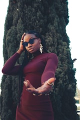 A person with long braided hair wearing a burgundy outfit and sunglasses stands in front of a tall, dense evergreen tree. They have one hand raised towards the camera and wear large, leaf-shaped earrings.