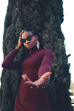 A person with long braided hair wearing a burgundy outfit and sunglasses stands in front of a tall, dense evergreen tree. They have one hand raised towards the camera and wear large, leaf-shaped earrings.