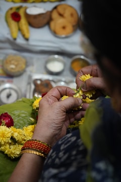 A person is crafting a yellow flower garland, with hands visible holding and arranging the flowers. The background includes various traditional items such as bananas, a coconut, snacks, and bowls of offerings, suggesting a religious or festive setting. The person is wearing colorful bangles and patterned clothing.