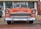 A classic orange car with chrome accents, parked on a street in front of a building. The car features a prominent front grille and round headlights. A small American flag is visible in the background.