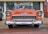 A classic orange car with chrome accents, parked on a street in front of a building. The car features a prominent front grille and round headlights. A small American flag is visible in the background.