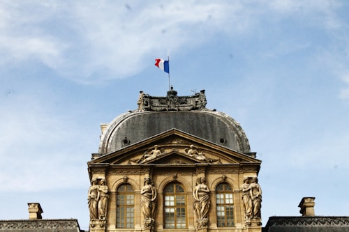 A grand architectural structure with ornate details, featuring a golden facade adorned with statues and intricate carvings. At the top, there is a tri-color flag prominently displayed against a clear blue sky. The building exudes an air of classical elegance and sophistication.