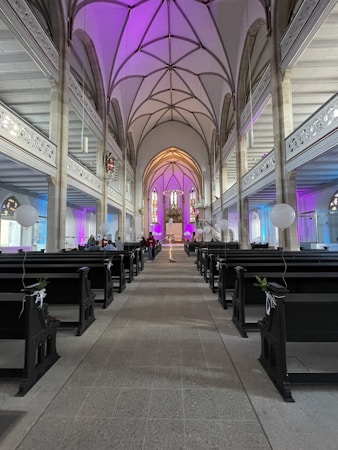 A spacious church interior with a high arched ceiling adorned with intricate designs. The aisle is flanked by dark wooden pews decorated with white ribbons and balloons. Soft purple and gold lighting enhances the ambiance, leading toward the altar at the far end, featuring stained glass windows.