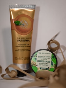 A tube of Satsuma-scented hand cream from The Body Shop stands beside a small circular container of Sugar Lip Scrub labeled as a vegan product. Both are surrounded by decorative curled paper. The hand cream features cold-pressed satsuma oil from Argentina, and the lip scrub is called Kindness & Pears, offering nourishing exfoliation.