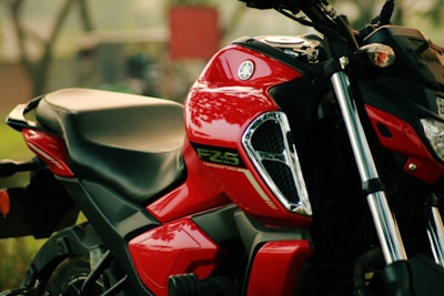 A close-up view of a red motorcycle with shiny chrome details on the side. The focus is on the tank and seat area, showing the logo and the model name 'FZ-S Version 3.0'. The background is blurred, with hints of greenery and an indistinct red object.
