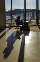 A person sits in an airport terminal, illuminated by sunlight streaming through large windows. The silhouette is cast prominently on the floor, capturing the tranquil ambiance of waiting. Their belongings are placed on the side, and the outside view reveals airport infrastructure.