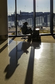 A person sits in an airport terminal, illuminated by sunlight streaming through large windows. The silhouette is cast prominently on the floor, capturing the tranquil ambiance of waiting. Their belongings are placed on the side, and the outside view reveals airport infrastructure.