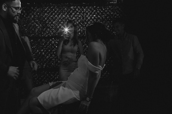 A group of people are socializing in a dimly lit environment, with one person capturing the moment using a smartphone flash. A woman in the foreground is wearing a strapless dress, engaging in a playful or dance pose, surrounded by others who appear to be enjoying themselves.