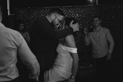 A cinematic black-and-white shot of wedding guests dancing joyfully in a dimly lit room.