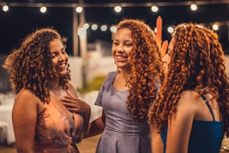Three women with curly hair are gathered together, smiling and engaging in conversation. The background is adorned with string lights, giving the scene a warm and festive atmosphere. They are dressed in colorful dresses.