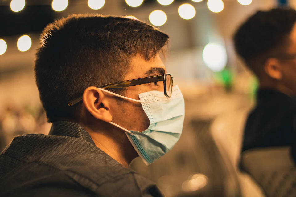 Man sits at a table wearing glasses and a face mask, partially obscured from view